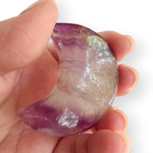 Hand holding Side view of a polished rainbow fluorite crescent moon displaying natural layers of purple and white fluorite.