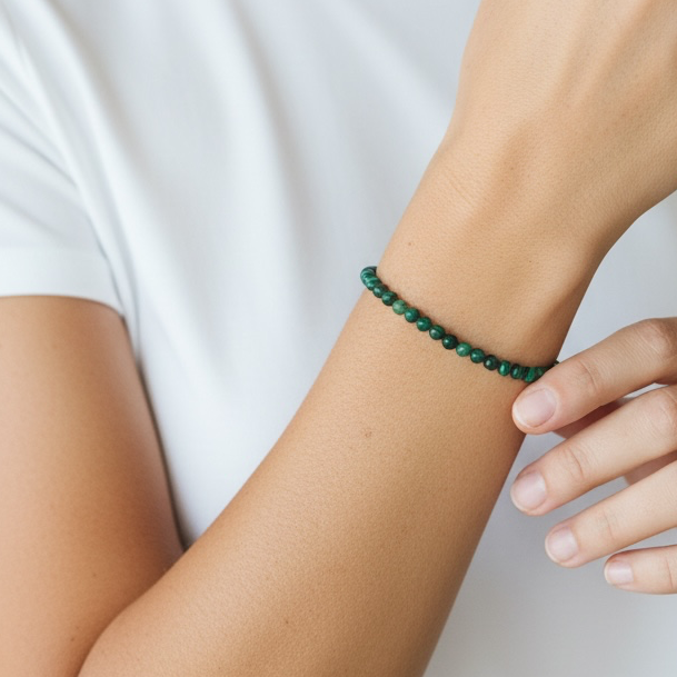 A close-up of a woman wearing a thin beaded moss agate bracelet in dark green, styled with a white t-shirt against a soft, light gray background.