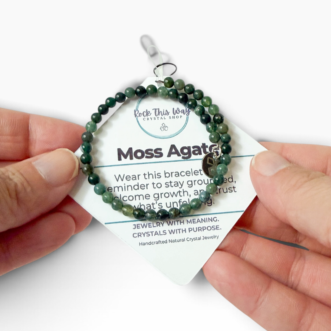 A person's hands holding a green moss agate beaded bracelet on a white display card. The card features the brand logo 'Earth This Way' and the text 'Moss Agate' along with a crystal affirmation.