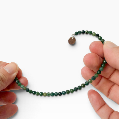 A beaded bracelet made of small green moss agate beads with a small charm, held between two hands against a plain white background.