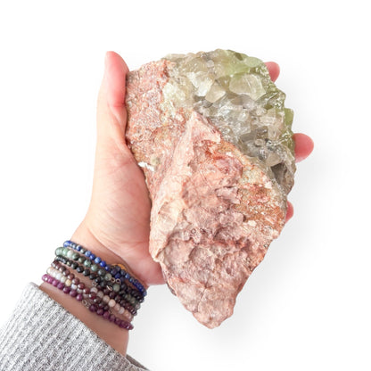 Hand holding large Green Calcite cluster to show size and rough base of red rock matrix.